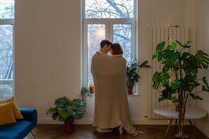 Rear view romantic couple wrapped in blanket looking out window at home. Family warming under plaid photo