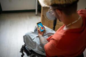 Disabled guy in wheelchair checking weather forecast in mobile app on smartphone at home. Heat wave photo
