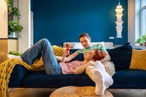 Loving couple resting on couch using smart phone apps. Wife showing interesting website to husband photo