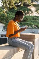 alegre africano americano mujer riendo mirando a móvil teléfono pantalla descansando teniendo bueno hora fuera de foto