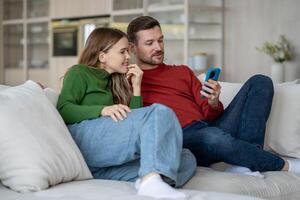 alegre casado Pareja sonriente, riendo sentado en sofá con teléfono hojeada en línea Internet contenido foto