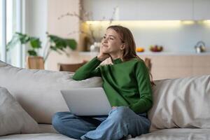 Thoughtful female remote worker relaxing on sofa with computer, distracted from business, thinking. photo