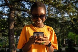 riendo africano americano mujer utilizando teléfono, felizmente sonriente mientras mirando a teléfono inteligente pantalla. foto