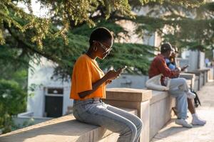 satisfecho optimista africano americano mujer con teléfono inteligente al aire libre sentado en parque en soleado clima foto