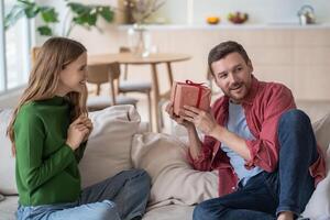 Amazed man, holding gift from loving wife, tries to guess contents of box sitting on couch at home. photo