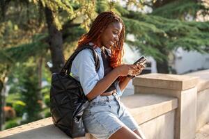 contento africano americano niña estudiante sonriente mira a pantalla teléfono tipo mensaje después Universidad al aire libre foto