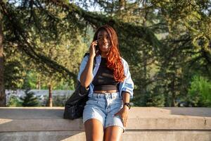 Pleased African American woman taking phone call while walking strolling in park in setting sun. photo