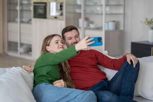 Joyful couple sitting on sofa, looking into smartphone camera, making selfie for sending to parents photo