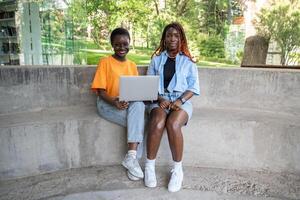 Happy contented black girls students sit with laptop near college library smiling looking at camera photo