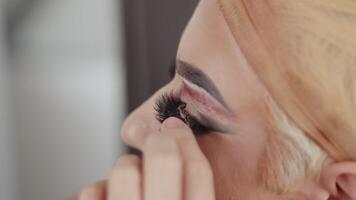 joven ejecutante en arrastrar reina maquillaje aplicando falso pestañas en su párpados en el vendaje habitación durante preparación para el actuación cerca arriba video