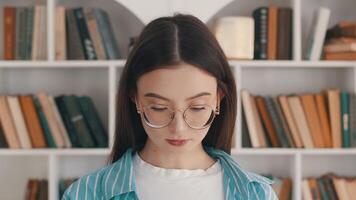 Portrait of a young caucasian woman in glasses raising her head, looking at the camera, and smiling showing her braces with the background of white bookcases with many books on the bookshelves video