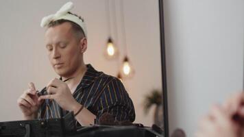Young man in drag queen makeup applying eyeshadows to his eyebrows using a brush and hand mirror in the dressing room during preparation for the performance video
