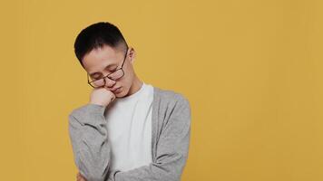 Young asian guy in glasses falls asleep standing, resting his head on his hand and then suddenly wakes up with a tired face on a yellow background in the studio video