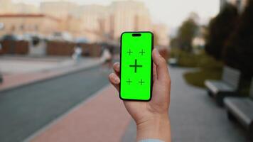 Close-up of a hand holding a smartphone with a mockup green screen with a sidewalk of a city street on a sunny day as a backdrop. Concept of a modern phone app mockup video