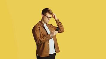 Young handsome caucasian man in glasses and casual clothes standing with a smartphone, scratching his head and looking perplexed on a yellow background in the studio video