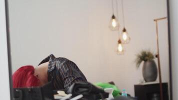 Young man in drag queen makeup putting on a long red wig on his head in front of the mirror and adjusting it in the dressing room during preparation for the performance video