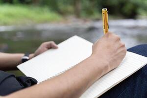 Young man takes notes in a notebook on nature. Man sits by a tree with his back to him, a pencil and a notebook in his hands. photo