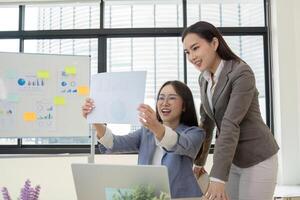 Two Female Accountant Collaborate on Financial Graphs in a Modern Office Setting with Large Windows and Natural Light photo