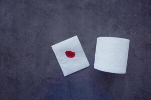 A blood spot is noticeable on a tissue next to a roll of toilet paper in the background photo