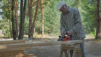 An elderly craftsman skillfully shapes a wooden plank in a serene forest setting. Surrounded by tall trees and natural light, he focuses on his work while using an electric planer to process wood. video