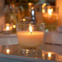 A single lit candle in a glass holder on a reflective surface against an out of focus background of bathroom toiletries photo