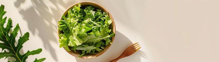 A wooden bowl of fresh green salad on a white table next to a wooden fork. photo