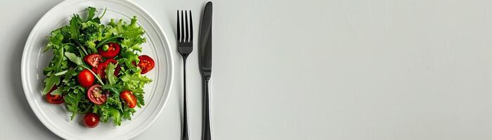 On a white plate is a salad of lettuce and red cherry tomatoes. Next to the plate is a black fork and knife. The background is a light grey. photo