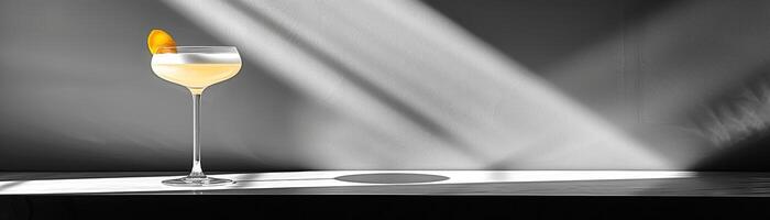A black and white photo of a martini glass with a orange peel on the rim sitting on a bar with light rays in the background.