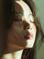 A close up portrait of a young woman with long dark hair and light makeup. She has her eyes closed and her face is turned slightly to the right. The light is shining on her face from the left side of photo