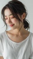 A photo of a young woman with long dark hair and a white shirt. She is smiling and looking down. Her hair is styled in a ponytail with loose waves.