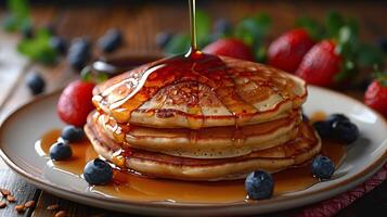 A stack of pancakes with blueberries and strawberries on the side and syrup being poured on top photo