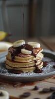A decadent stack of pancakes topped with sliced bananas and drizzled in chocolate syrup. photo