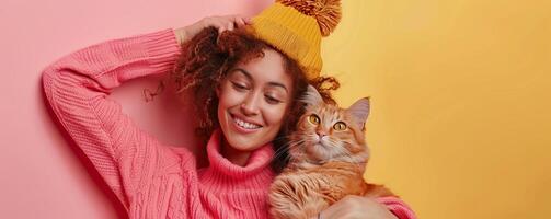 Young woman in a pink sweater and yellow hat smiling while holding a ginger cat against a pink and yellow background. photo