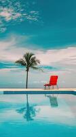 Serene poolside scene with a single red lounge chair, palm tree, and clear blue sky reflecting on the water. Perfect for travel and relaxation themes. photo