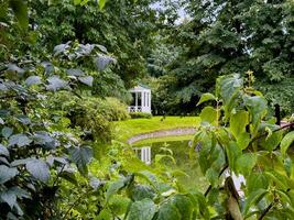 A peaceful garden scene featuring a charming white gazebo reflecting in a tranquil pond, surrounded by lush greenery. The serene atmosphere is enhanced by the soft lighting and natural beauty, making photo