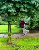 A woman with whimsical fairy wings playfully dances under the shade of a large tree in a vibrant green park. The lush foliage and bright grass create a magical and enchanting atmosphere, as if photo