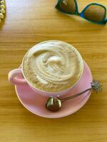 A high-angle shot of a freshly brewed cappuccino in a pink cup and saucer, served with a unique tropical-themed spoon on a wooden table. The warm, inviting scene includes sunglasses in the background, suggesting a relaxing moment photo