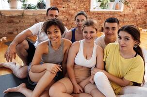 Multiracial smiling people sit on mats after doing yoga at fitness club. Group of young athletes of different age and nationalities relax and rest after physical exercises in the gym. photo
