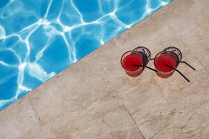 Two glasses with a tasty red cocktail with ice. Delicious fresh drink with straws by the pool. A sunny day and the reflection of rays in the blue water in background. photo