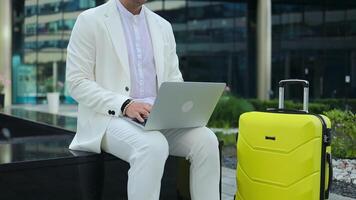Focused young businessman corporate worker investor with suitcase sitting near business centre waiting, working remote on laptop video