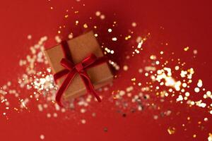Close up of a present box on a red background. Kraft paper gift with red velvet ribbon and bow. Bright backdrop with silver sparkles and gold round confetti. photo
