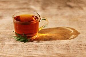 A transparent cup of delicious strong black tea with a cannabis leaf. A glass cup and hemp on a wooden table. A healthy plant-based organic drink. photo