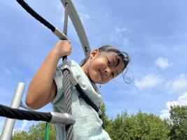 A happy young Asian little girl bravely climbing on a rope in the playground. Childhood concept. Down view photo