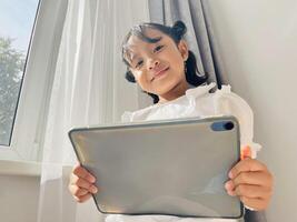 Asian little child standing near the window and holding mobile tablet smiling at the camera. Gadget addicted concept. Low angel photo