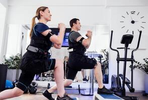 Sport couple in ems suits is engaged in physical activity in fitness club or gym. Man and woman with clenched hands doing forward lunges with step platforms. photo