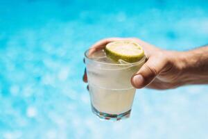 Young male hand holds a glass of a chilled drink with ice cubes. White cocktail with a lemon slice. Sunny day and reflection of rays in the blue pool water on background. photo