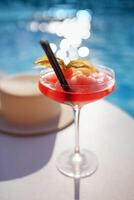 A red drink in an elegant glass with a long stem on the side of the pool. An alcoholic ice drink decorated with a flower on a background of turquoise water. photo