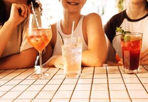 Close-up of three charming girls drinking chilled cocktails in a cafe. Glasses with delicious fruit drinks and ice cubes. Smiling friends relax and drink lemonade in a restaurant. photo
