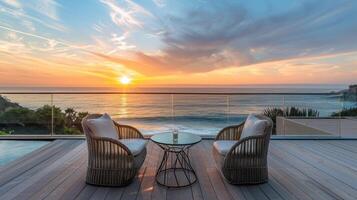 Two round wicker chairs with cushions and a small wicker table with a plant, all set on a wooden deck overlooking a stunning ocean view. photo