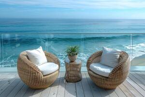 Two round wicker chairs with cushions and a small wicker table with a plant, all set on a wooden deck overlooking a stunning ocean view. photo